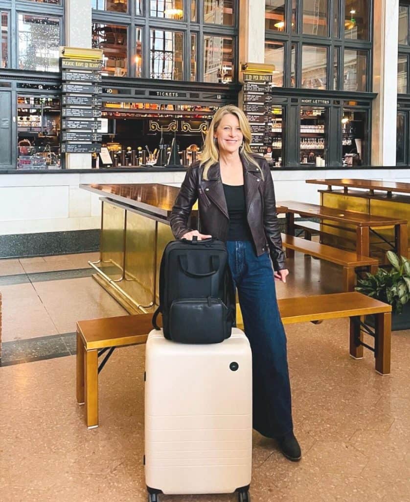 Susan Heinrich stands in Denver's Union Station with Monos luggage - the Monos Medium Check-In and the Monos Metro backpack.