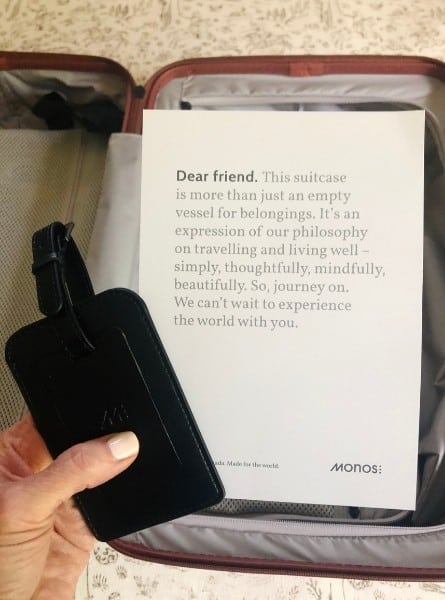A Monos suitcase with a hand holding a message about the meaning of travel