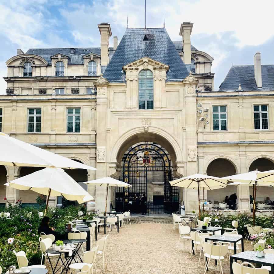 The Musee Carnavalet in Le Marais, Paris. The striking beige stone exterior is pictured with pitched slate roofs. The courtyard garden is in the foreground with tables, umbrellas and greenery.