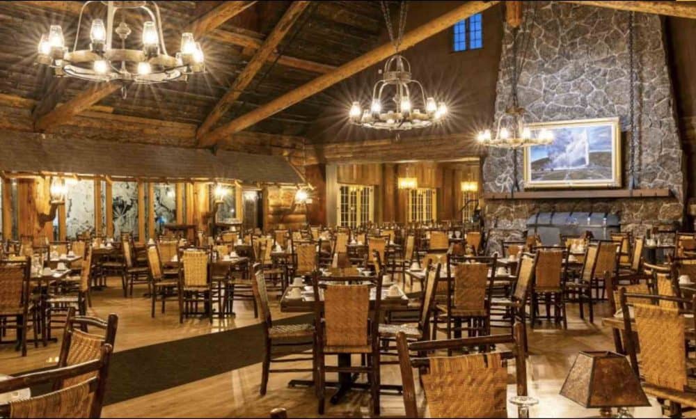 A historic wood beamed dining room at a national park inn. Wood tables an chairs, a stone fireplace and rustic chdeliers are visible.