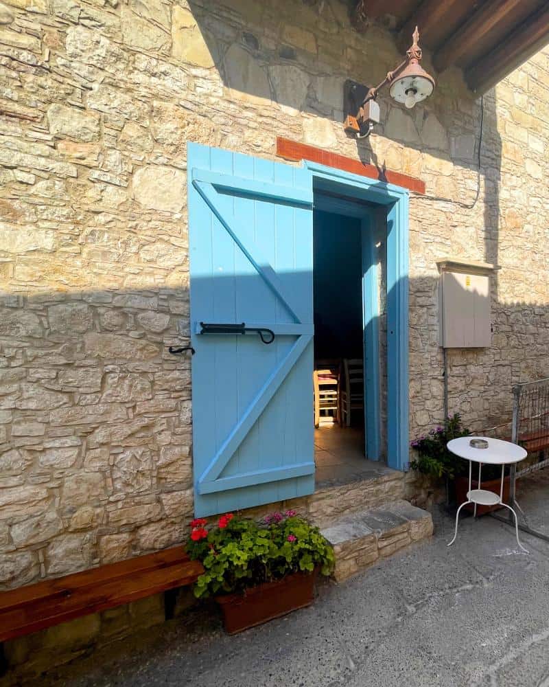 A turquoise door is open. It's set in a stone wall. A small white table, bench and red flower planter are also visible.