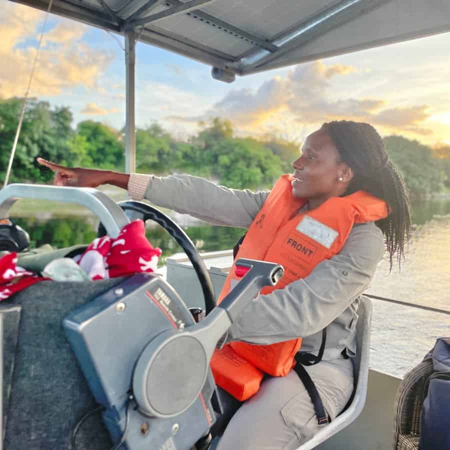 Chobe Game Lodge Guide, Oriah Nthobatsang, points to something out of view on the Chobe River. She is a seated at the wheel of a riverboat wearing a grey jumpsuit and am orange life jacket.
