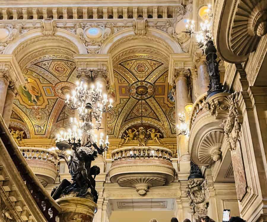Looking up the stairs at the ceiling of the Palais Garnier in Paris, with stunning Belle Epoque architecture and decorative light fixtures.