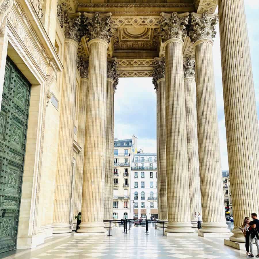 The entrance to the Pantheon Paris, a historic monument in Paris's Latin Quarter which now functions as a civic building. Pictured are enormous Corinthian-style pillars, a marble entry floor and a towering green door.