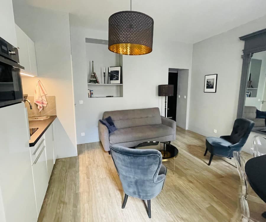 A Paris Airbnb in the 2nd arrondissement. Pictured is an open room with a grey sofa and two armchairs. There is also a dining table with chairs. On the left of the photo is a kitchenette.