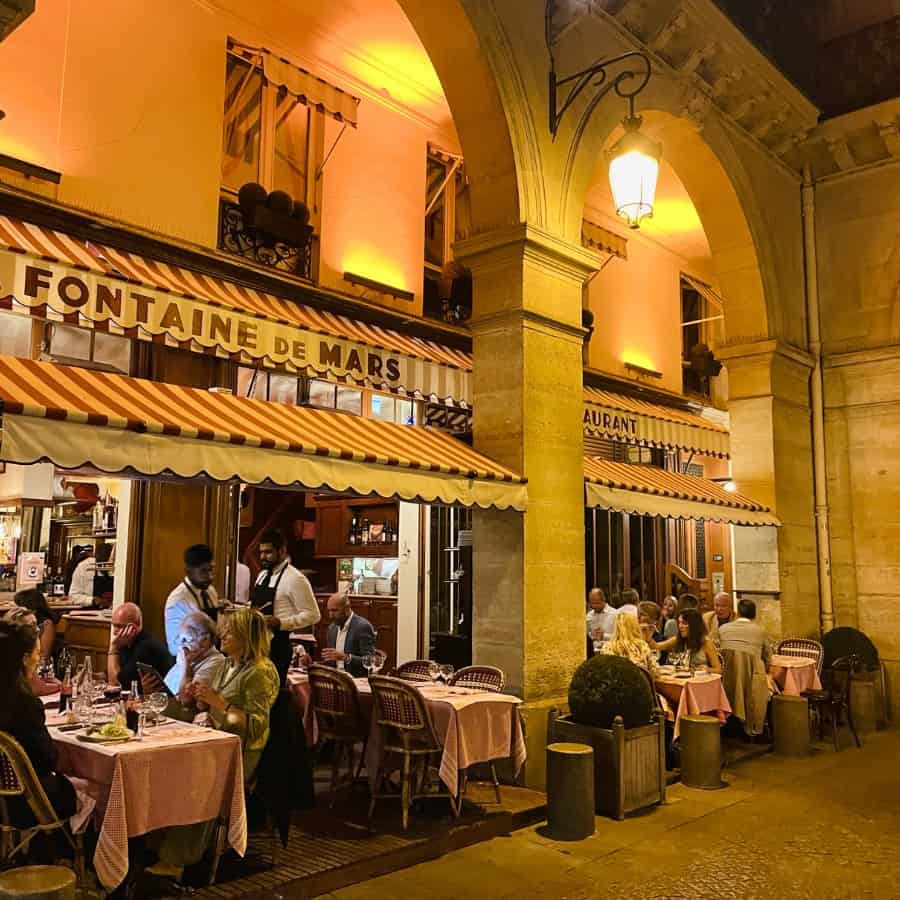 La Fontaine de Mars, a popular Paris restaurant in the 7th arrondissement. Pictured is a golden lit terrace in evening with a pretty pink striped awning.