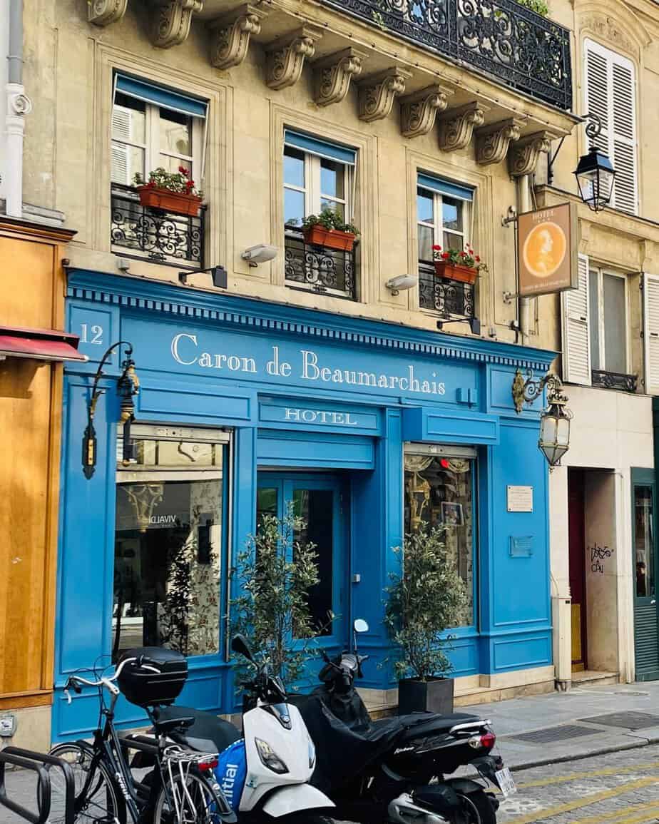 A colorful and charming exterior to a small hotel in Paris. The entrance is flanked in blue trim and lanterns, and above that are three traditional windows with red flowers in flower boxes.