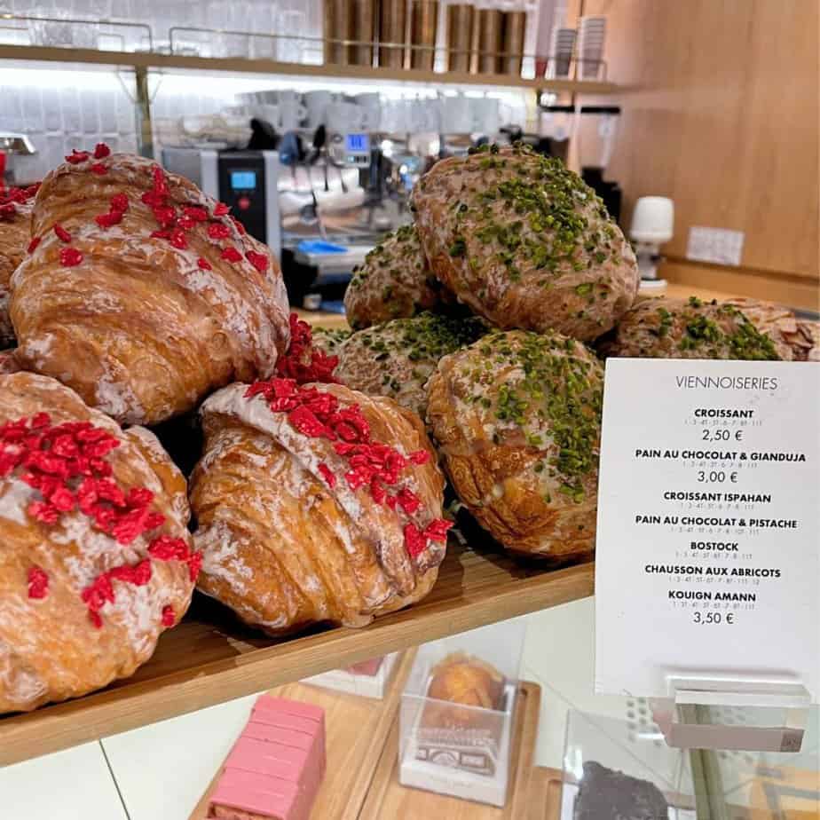 Colorful croissants sit atop a counter in a bakery. They have varying colors as decoration; some have rose sprinkles and the others have green. In front of that is a price list with the title vienoisserie at the top and a list of the pastries on offer.