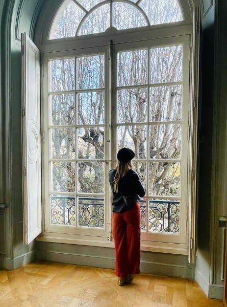 A woman stands looking out a window in the Rodin Museum in Paris. She is wearing red flowy pants, a black leather jacket, and a black beret.