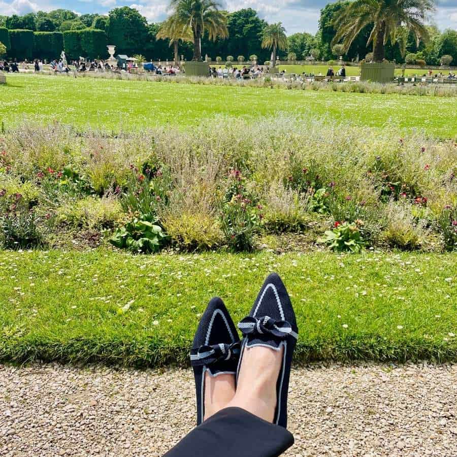 A pair of feet propped up in blue pointed loafers are pictured in a Paris park. Beyond is grass, gardens and trees.