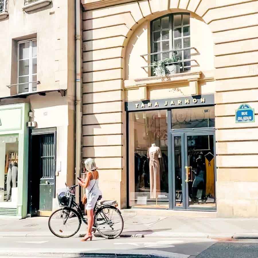 A woman on a bike in a dress in heels looks chic outside of a Tara Jarmon store in Paris