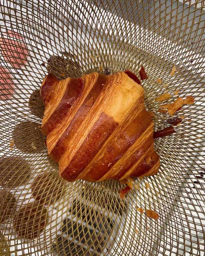 A single butter croissant sits in a pretty wire basket at a bakery in Paris. A few cookies are slightly visible below. It is a golden bronze color and there are flakes surrounding it.