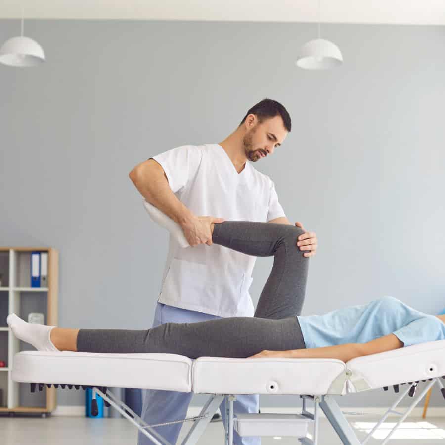 A woman wearing grey tights and a t-shirt lies on a therapy table and a therapist holds one of her legs at the knee and ankle