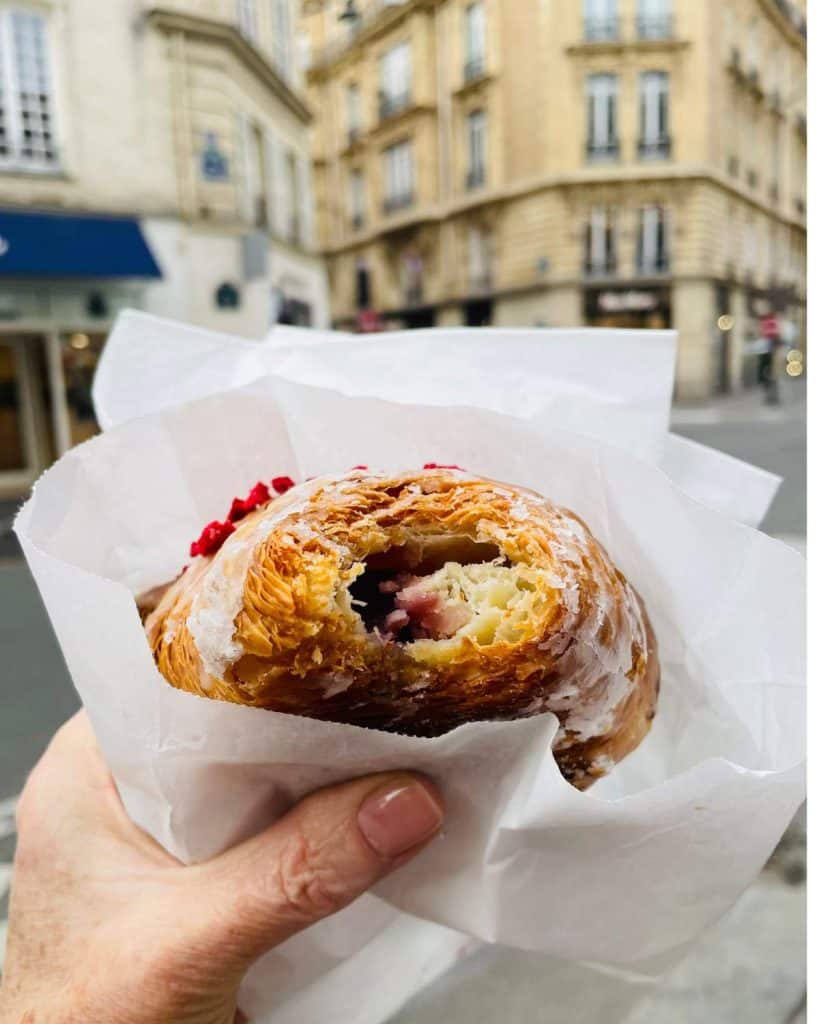 A croissant is being held out by a hand on a Paris street with Haussmann buildings beyond. It has a bite missing from the end. It has a pink-colored filling and rose decorative sprinkles on top.