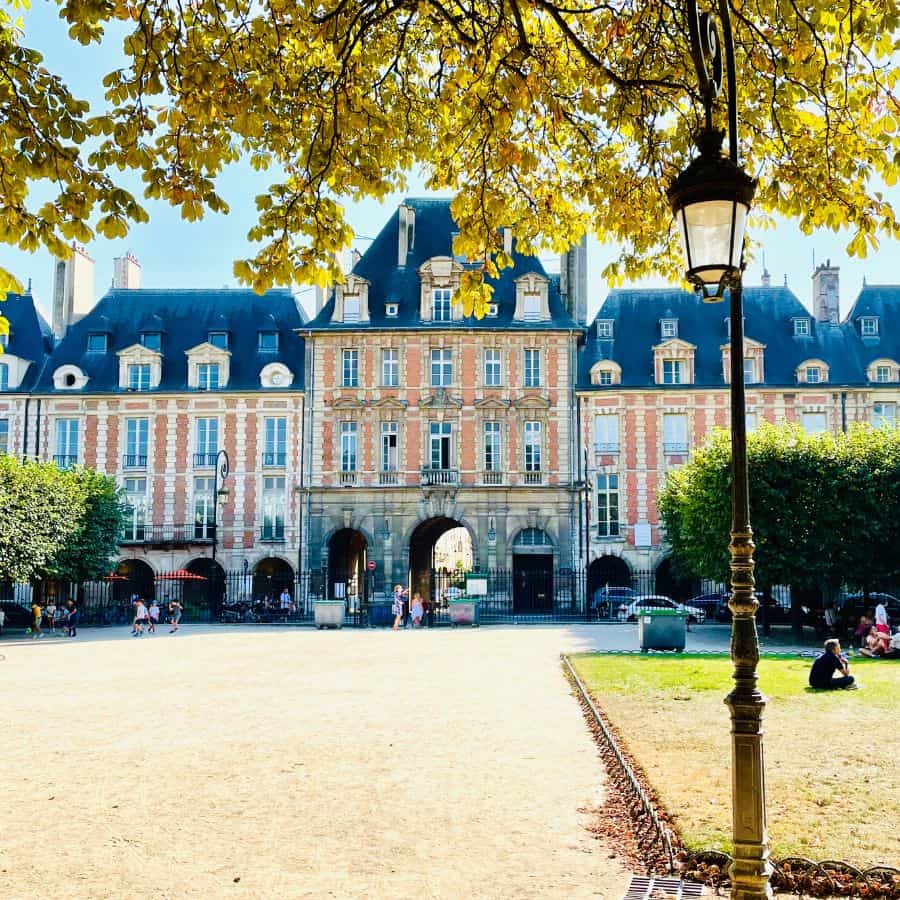 Place des Vosges Paris. A view of the park with an old lamp post on the right. This is Paris's oldest square inaugurated in 1612. Beyond the park are the striking architecture of identical house fronts: red brick with stone accents and pitched slate roofs, over vaulted arcades.