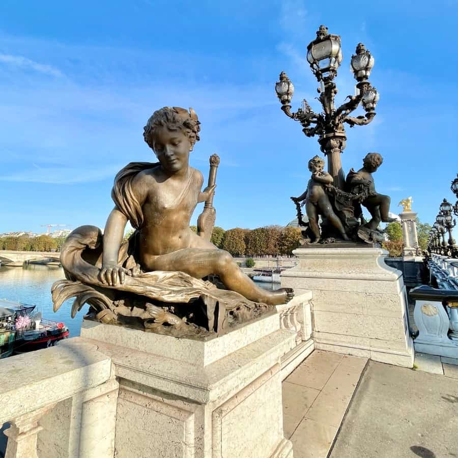 A beautiful sculpture of a nymph and striking Art Nouveau lamps at the edge of the Pont Alexandre III bridge in Paris. A little of the blue of the River Seine is visible beyond. This The Beaux-Arts style bridge is a Paris gem worth spending some time to admire.