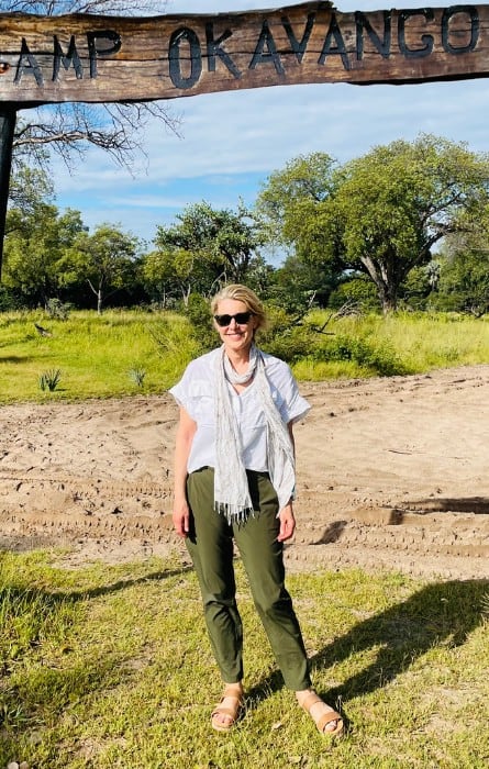 Susan Heinrich stands in front of a sign that says Camp Okavango at a safari lodge in Botswana. Her safari outfit is green pants, a white shirt, a cotton scarf and sandals.