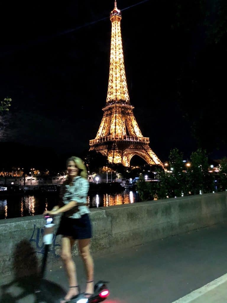 Susan Heinrich riding a scooter past the Eiffel Tower at night