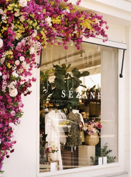 A window into the Grand Appartement Sézane store in Paris with clothing displayed in the window and an arch of pink, white and gold flowers overtop.