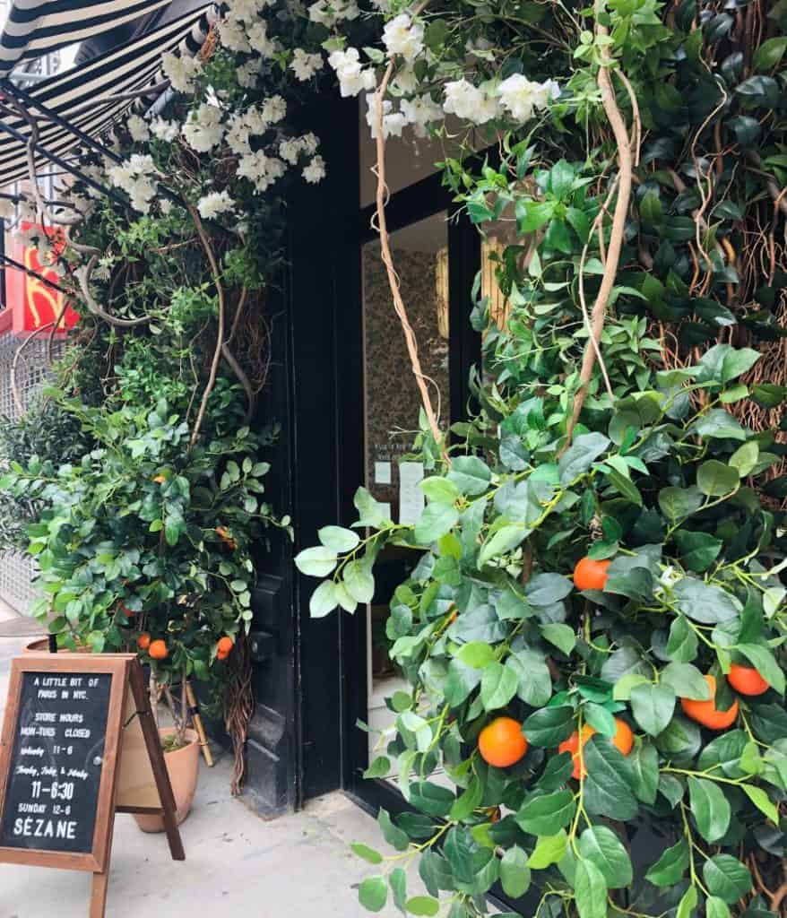 The exterior of the Sézane store in New York City in summer, with an archway of white flowers and bush with oranges. A easel-style sign lists store hours.