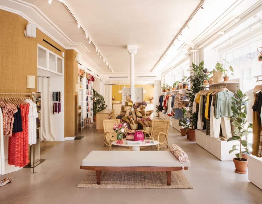 Inside the Sézane store, Le Grand Appartment, in Paris. Pictured is an poen airy space with benches in the center and racks of clothing displated on either side including trousers, blazers, sweaters and blouses. Items are in a range of colors.