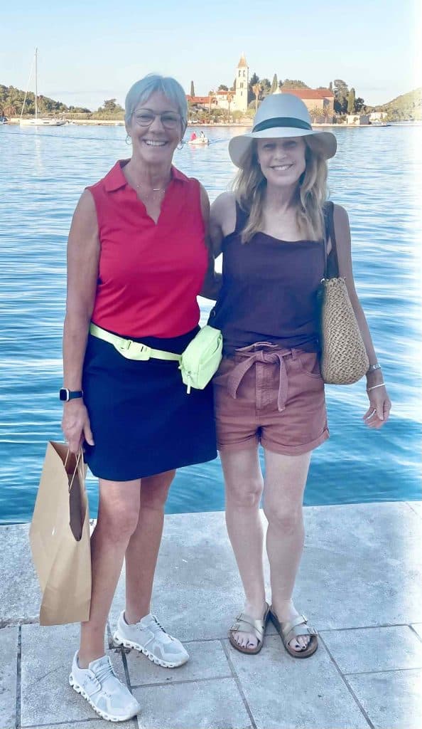 Two women stand together on a cement walkway in front of the sea.