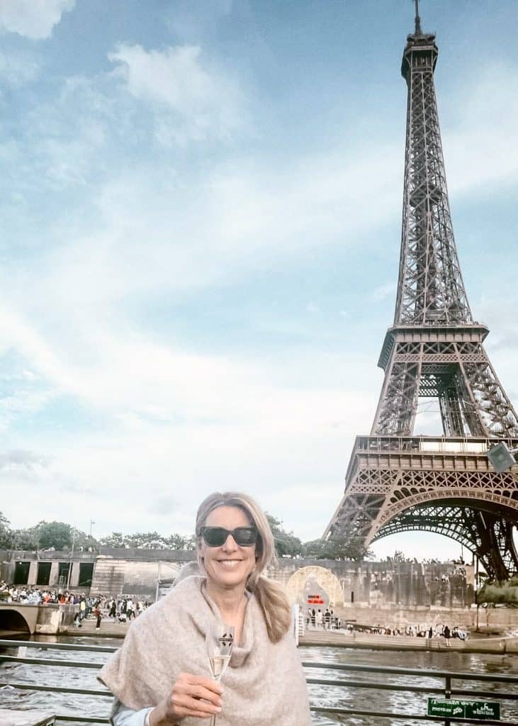 a woman stands holding with glass of champagne on a boat with the river and Eiffel Tower in the background