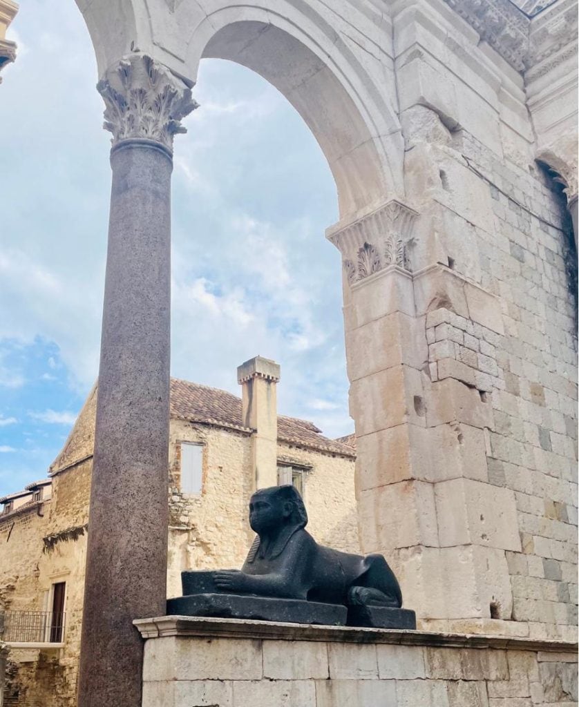 A sphinx sits in the ruins of the Palace of Diocletian in Split, Croatia. Intact arches and columns are visible.