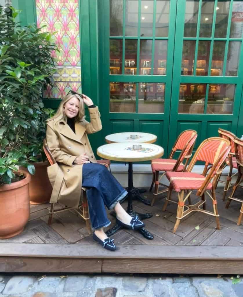 A woman sits on a Parisian terrace wearing a trench coat jeans and loafers.