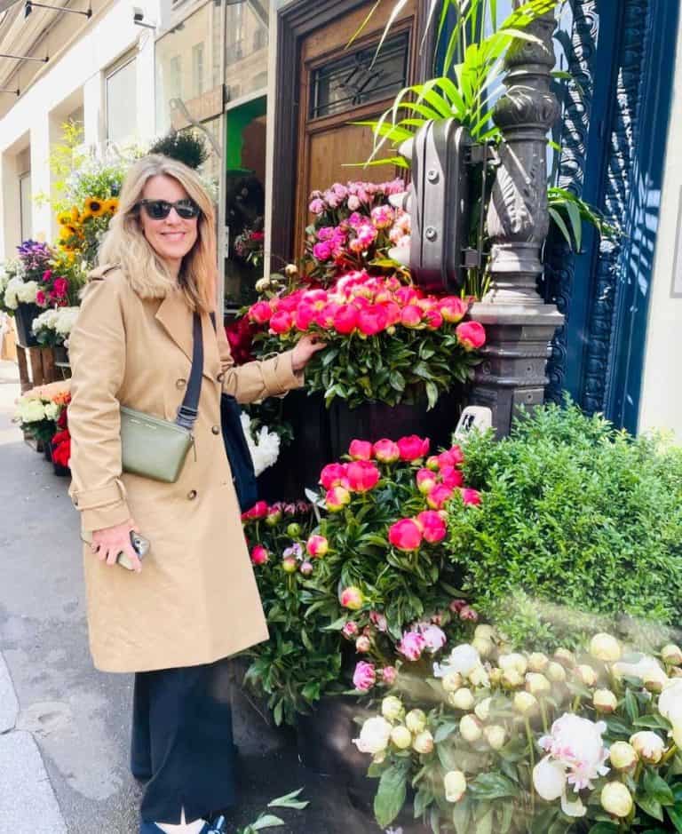 A woman stands in front of a Paris flower stand in spring. She is wearing a trenchcoat, dark pants and loafers and a sling bag.