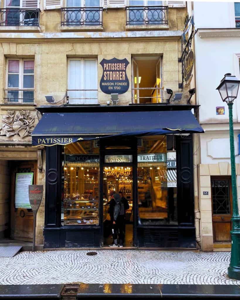 A Paris patisserie entrance has a blue awning and an open blue door. A few people are visible just inside the door. The street is cobblestone.