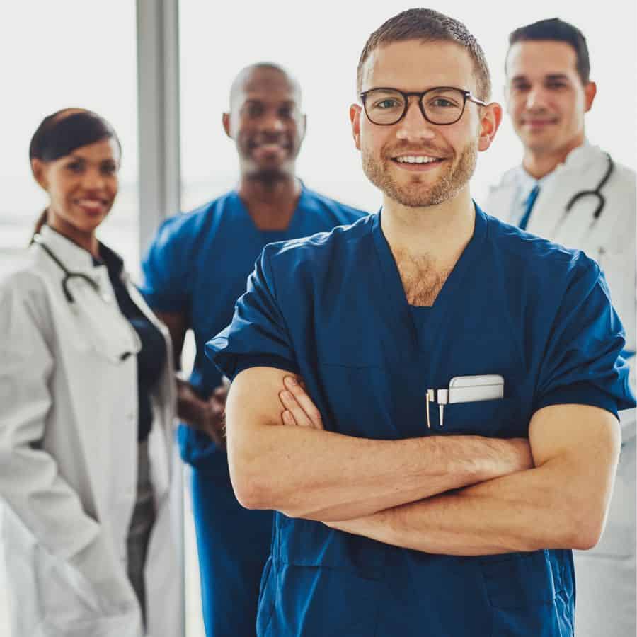 Four doctors stand together, three are men and one is a woman. Two are wearing white lab coats and two are dressed in blue scrubs