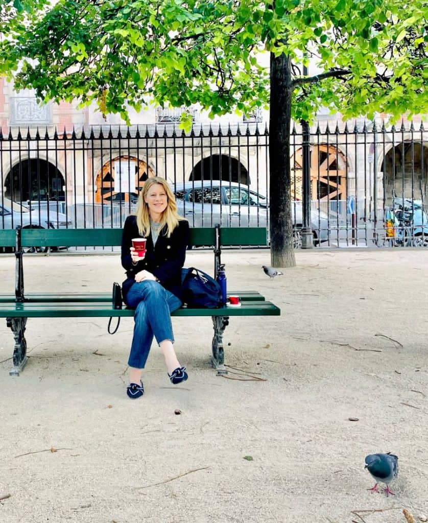 A woman sits on a bench in a Parisian park. She is wearing a blazer, jeans and loafers and holding a coffee cup.