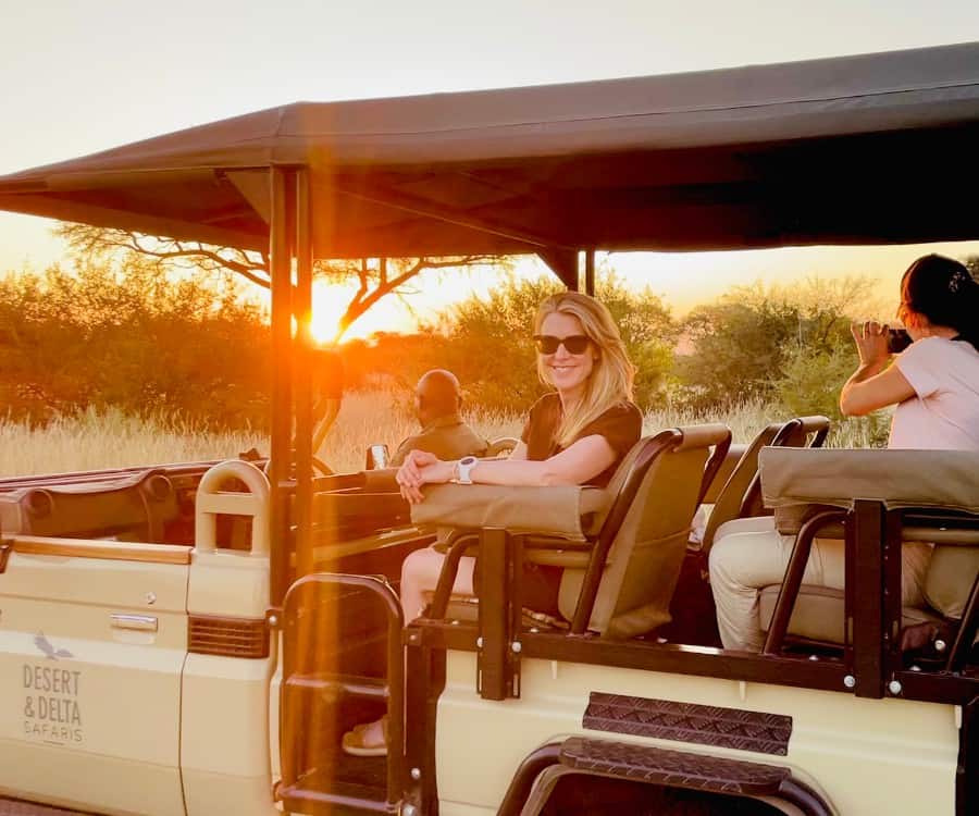 Susan Heinrich sits in a safari truck with another guest and her guide, Ollie, with the sun setting beyond.