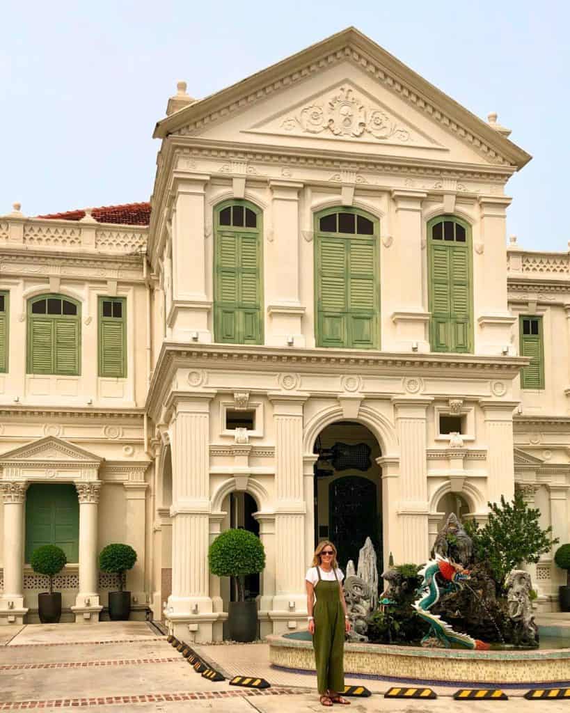 Susan Heinrich stands in front of The Edison Hotel in Penang Malaysia. It is a white Colonial-Style mansion dating to the early 1900s. It has a peaked roof and green shutters.