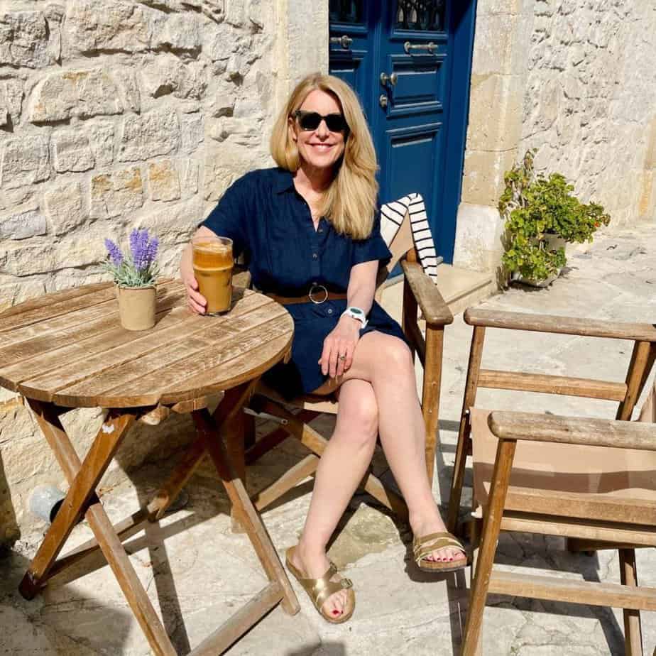 Susan Heinrich sits at a wooden café table in Cyprus, Europe on a cobblestone laneway. She is wearing a navy short-sleeved shirtdress and gold Birkenstock sandals and sunglasses. There is an iced coffee on the table as well as a small pot of lavender.