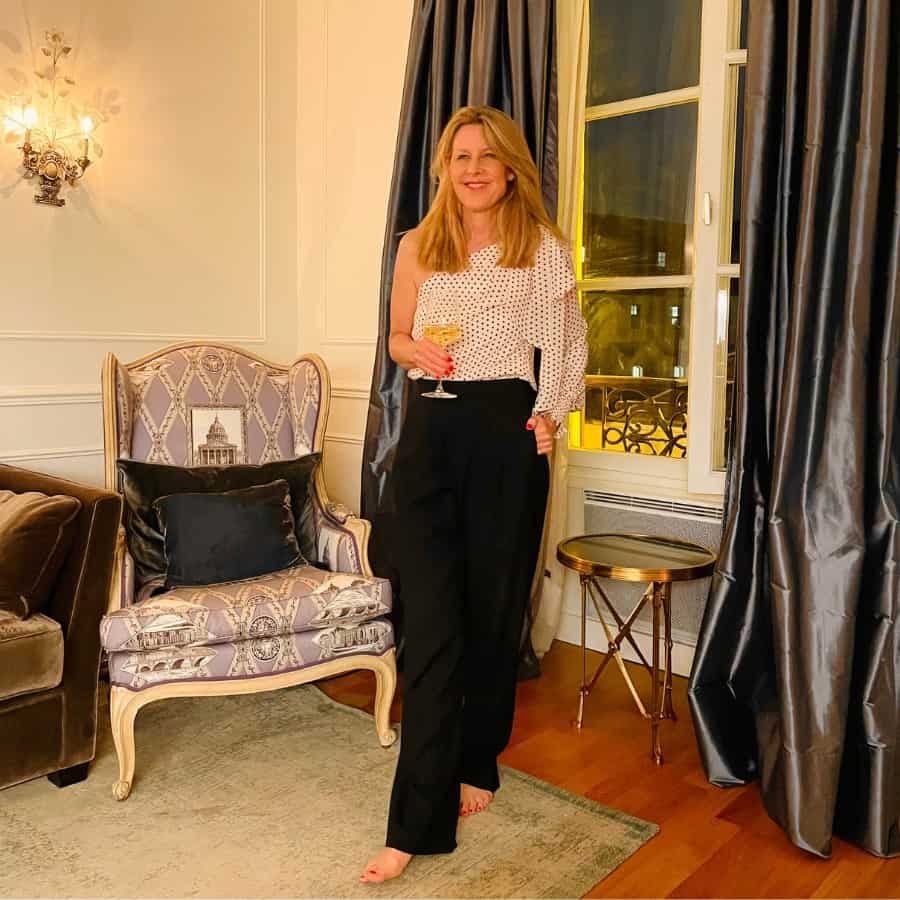 Susan Heinrich stands in a glamorous Paris apartment dressed in black trousers and a polka dot one-sleeved blouse.