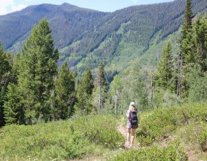 Susan Heinrich is pictured in the distance hiking on a trails at Vail. There are pine trees and green hills beyond her.