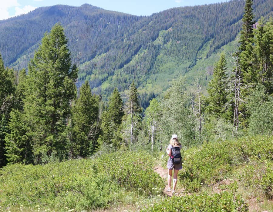 Susan Heinrich is pictured in the distance hiking on a trails at Vail. There are pine trees and green hills beyond her.