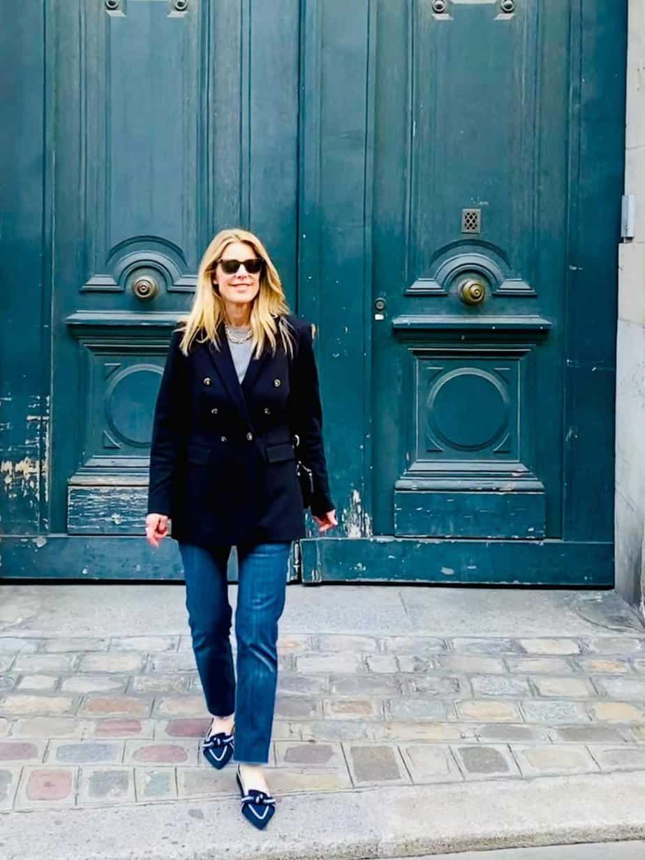 Susan Heinrich stands in front of a large blue door on cobblestones in the Marais area of Paris. She is wearing a black blazer, jeans and blue ballet flats with a bow.