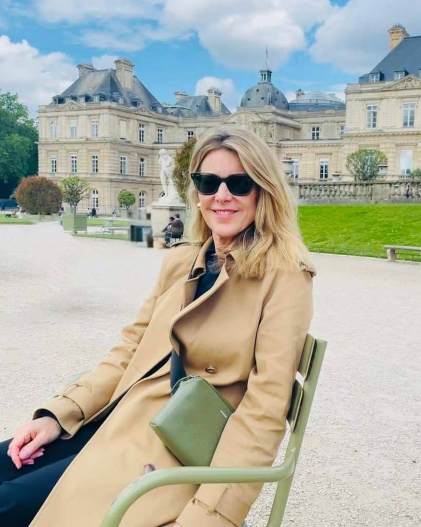 A woman wearing a beige trenchcoat and with sunglasses on, sits in a park with a gorgeous old building beyond her. It is an Italian-Renaissance palace.