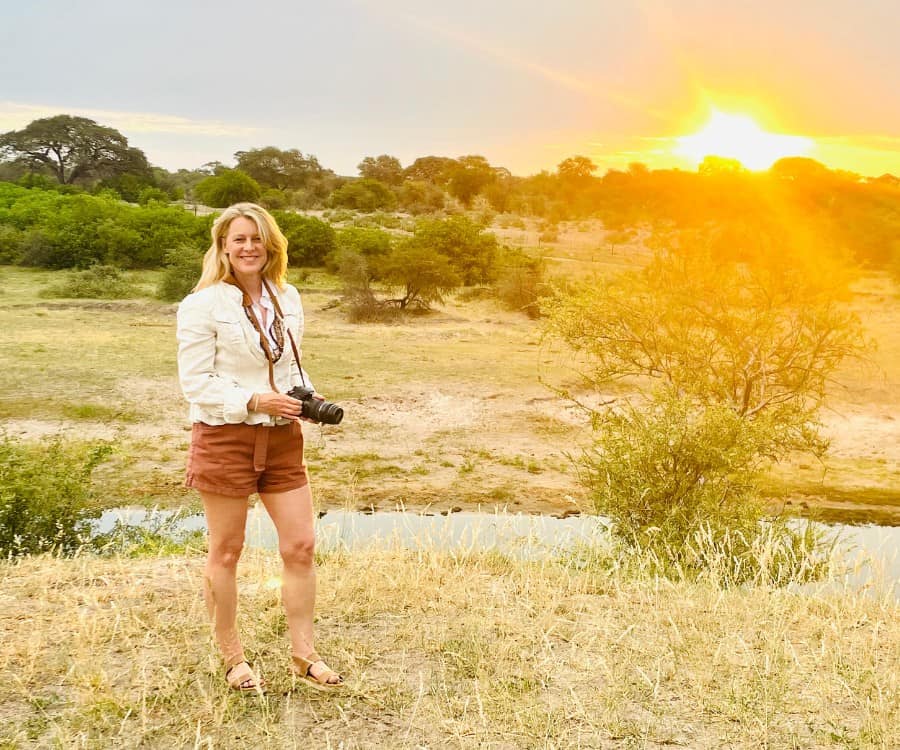 Susan Heinrich stands with her camera with the sunset beyond while on safari in Botswana's Makgadikgadi Pans National Park. She wears part of her safari wardrobe: rust shorts and a beige linen utility jacket.