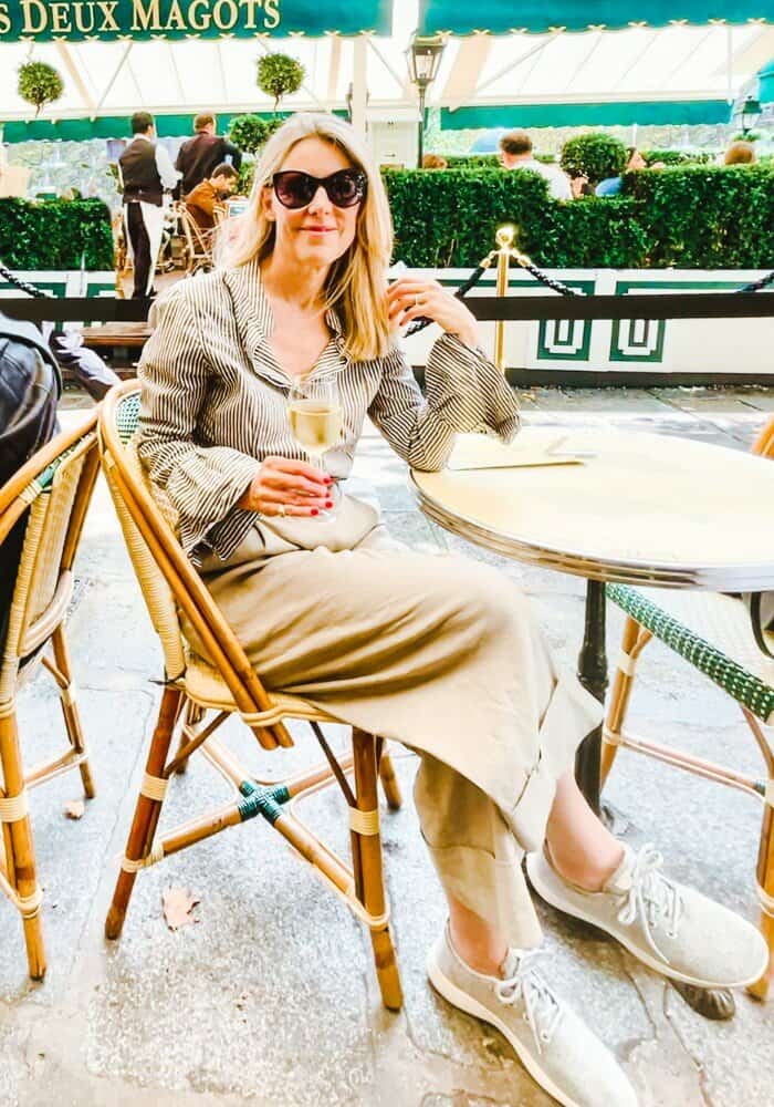 Susan Heinrich sits on the patio at Les Deux Magots in Paris in fall. She is wearing sunglasses, light trousers, a striped button up shirt and sneakers.