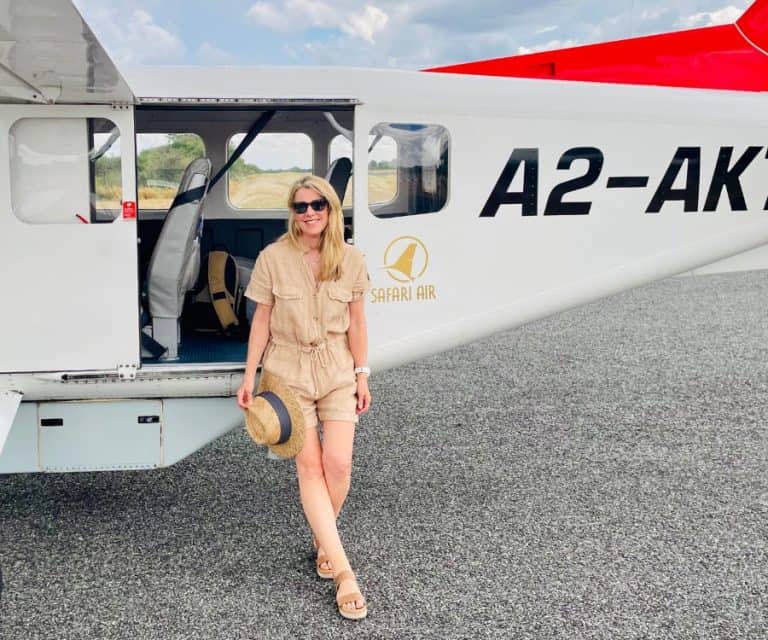 Susan Heinrich leans against the side of a small 6-seater airplane while on safari in Botswana. She wears a beige linen shorts romper and is holding a hat.