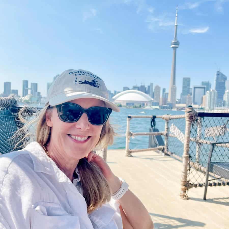Susan Heinrich sits on a water taxi in the Toronto Harbor with the city skyview beyond. She is smiling and wearing a white shirt and baseball hat.
