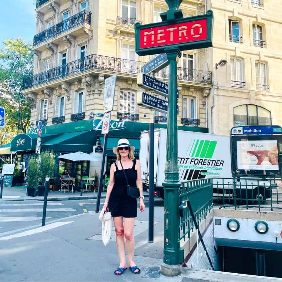 Susan Heinrich on a solo trip to Paris, standing in front of a Paris Metro sign with a pretty Haussmann building beyond