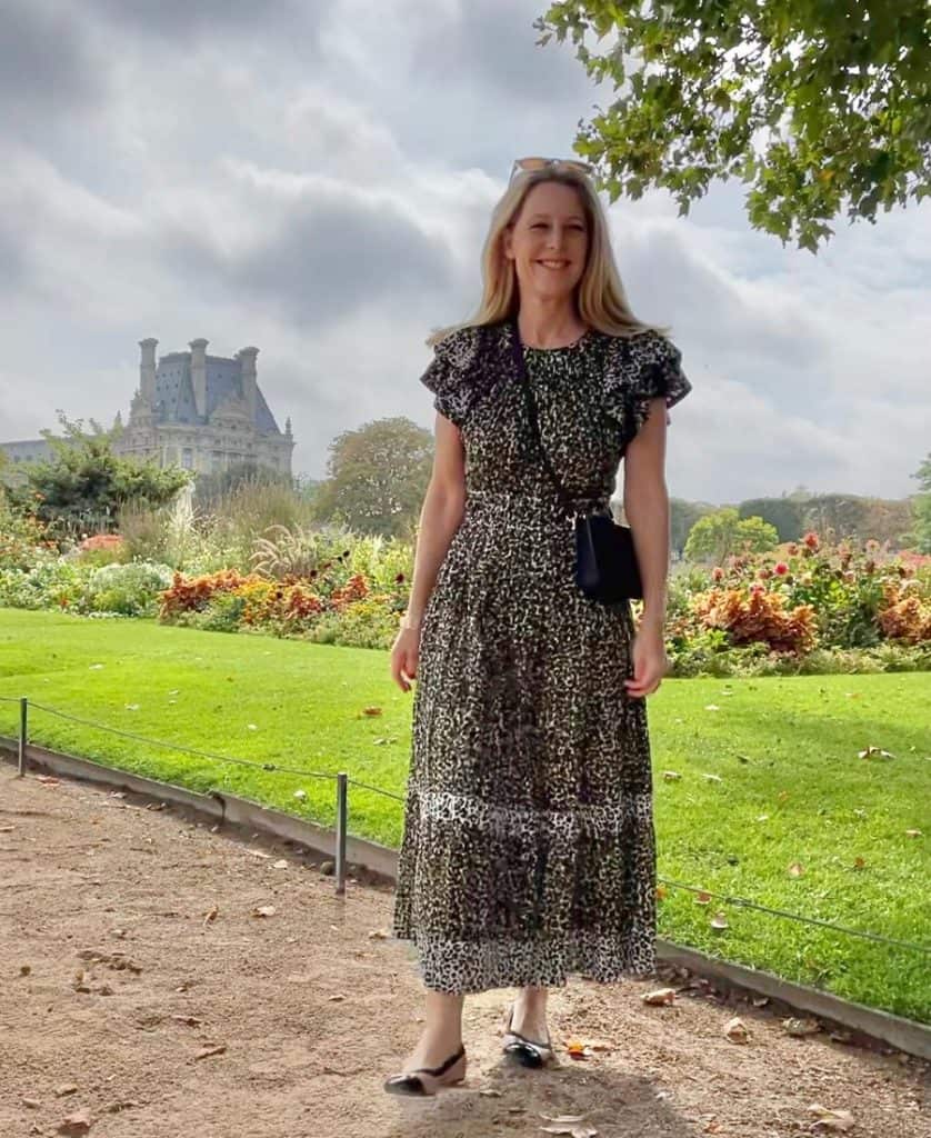 A woman stands in a large Parisian garden wearing a patterned dress and ballet flats