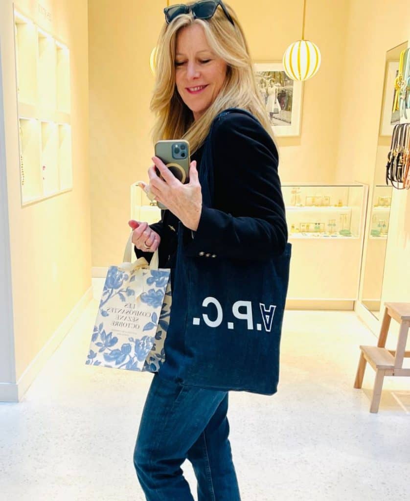 A blonde woman stands in a store with a navy tote bag over her shoulder withe the letters A.P.C. She also holds a small floral shopping bag