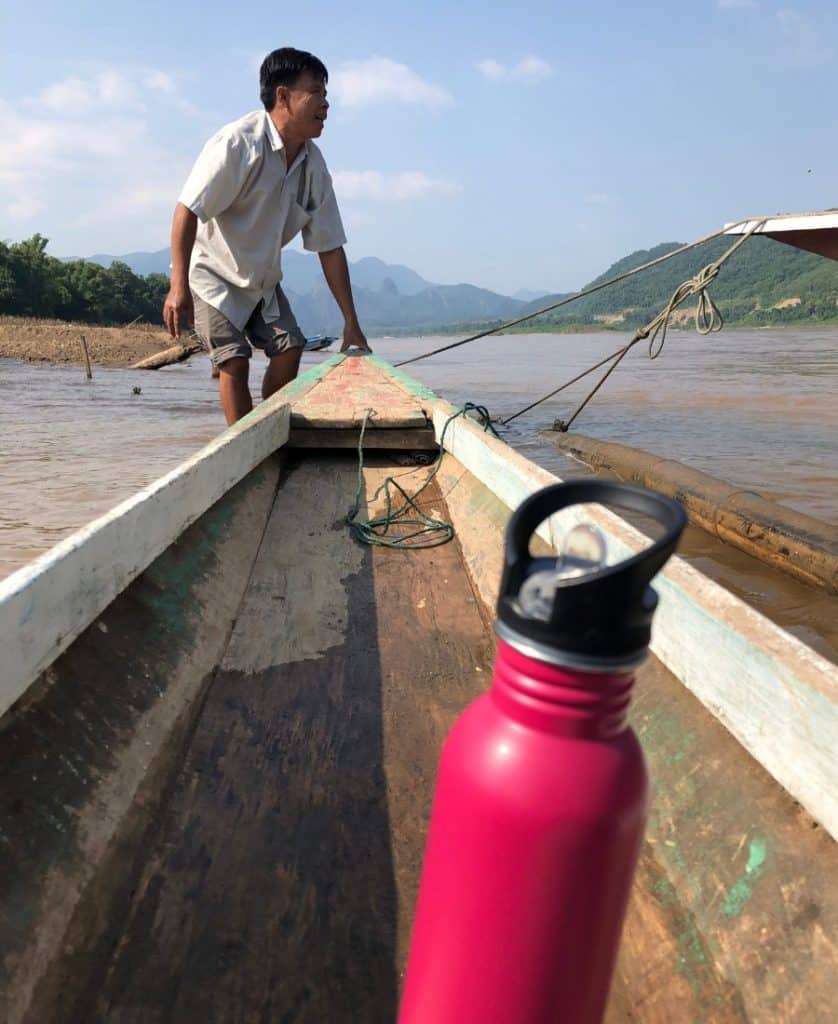A long-tail boat is pictured on the Mekong River in Luang Prabang, Laos. A pink reusable water bottle is pictured in the foreground and a guide is standing in the water holding the tip of the boat.