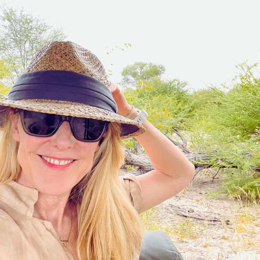 Susan Heinrich wears a favorite travel hat while sitting in a safari jeep on an Africa safari in Botswana. She is also wearing sunglasses and a beige linen romper. She has her hand raised to holding the back of her hat.